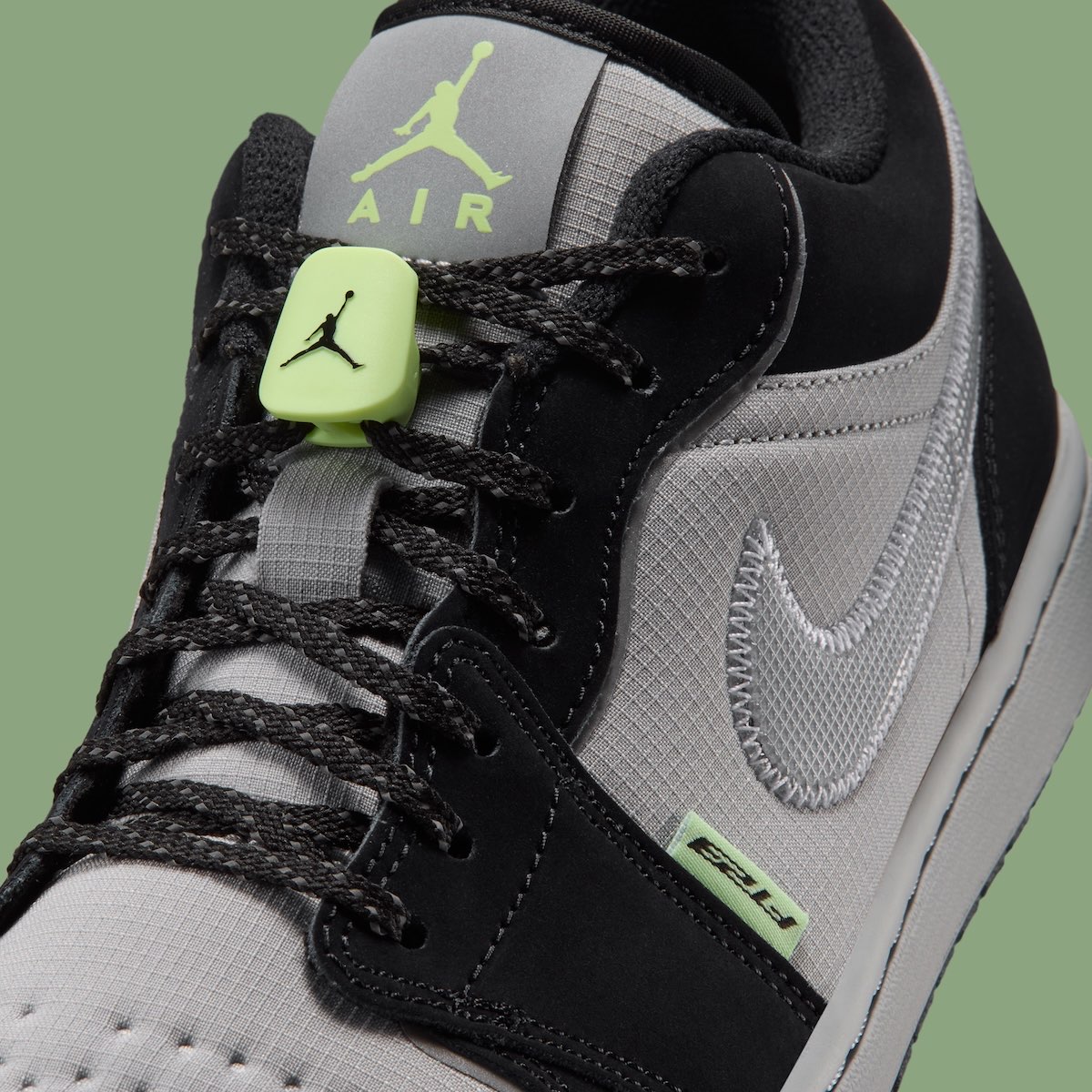 Detalle del Air Jordan 1 Low Light Smoke Grey Barely Volt con la traba de cordones verde flúor con el Jumpman, la lengüeta con el logo Jumpman Air en verde, los cordones negros tipo trekking y el Swoosh plateado cosido en zigzag sobre el ripstop