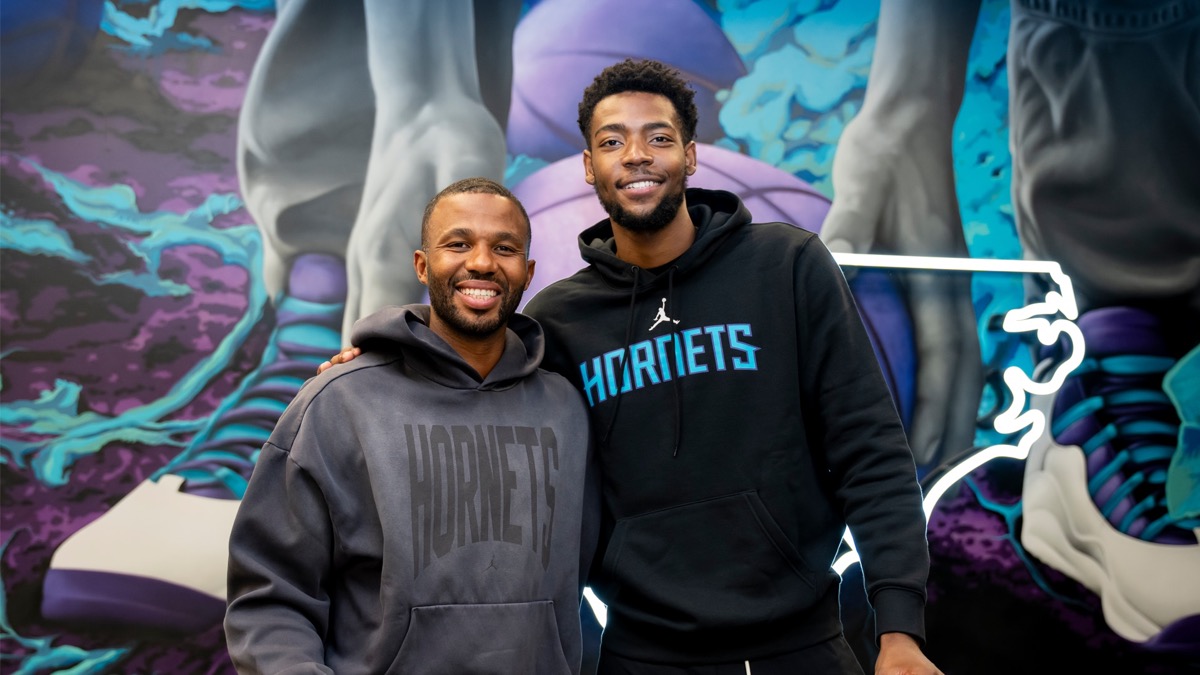 Brandon Miller, con un buzo negro de los Hornets, abrazado con un directivo del club frente a un mural con los colores de Charlotte