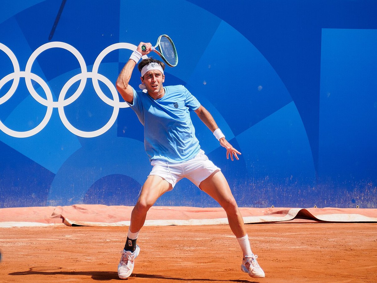 Foto de archivo: Tomás Martín Etcheverry, con remera celeste y vincha blanca, prepara un drive sobre polvo de ladrillo en los Juegos Olímpicos de París 2024, con los anillos olímpicos en la pared azul del fondo