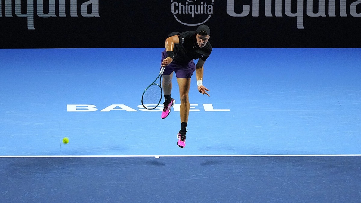Foto de archivo: Jakub Mensik, de negro y con zapatillas rosas, salta para sacar en la cancha azul del torneo de Basilea 2025, con la palabra Basel pintada en el fondo