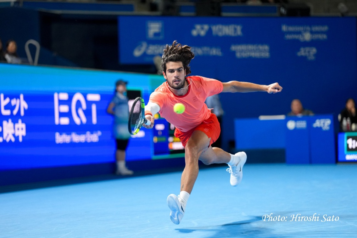Carlos Alcaraz, con remera coral y short rojo, se estira para pegar un revés frente a los carteles azules del Japan Open en el Ariake Coliseum