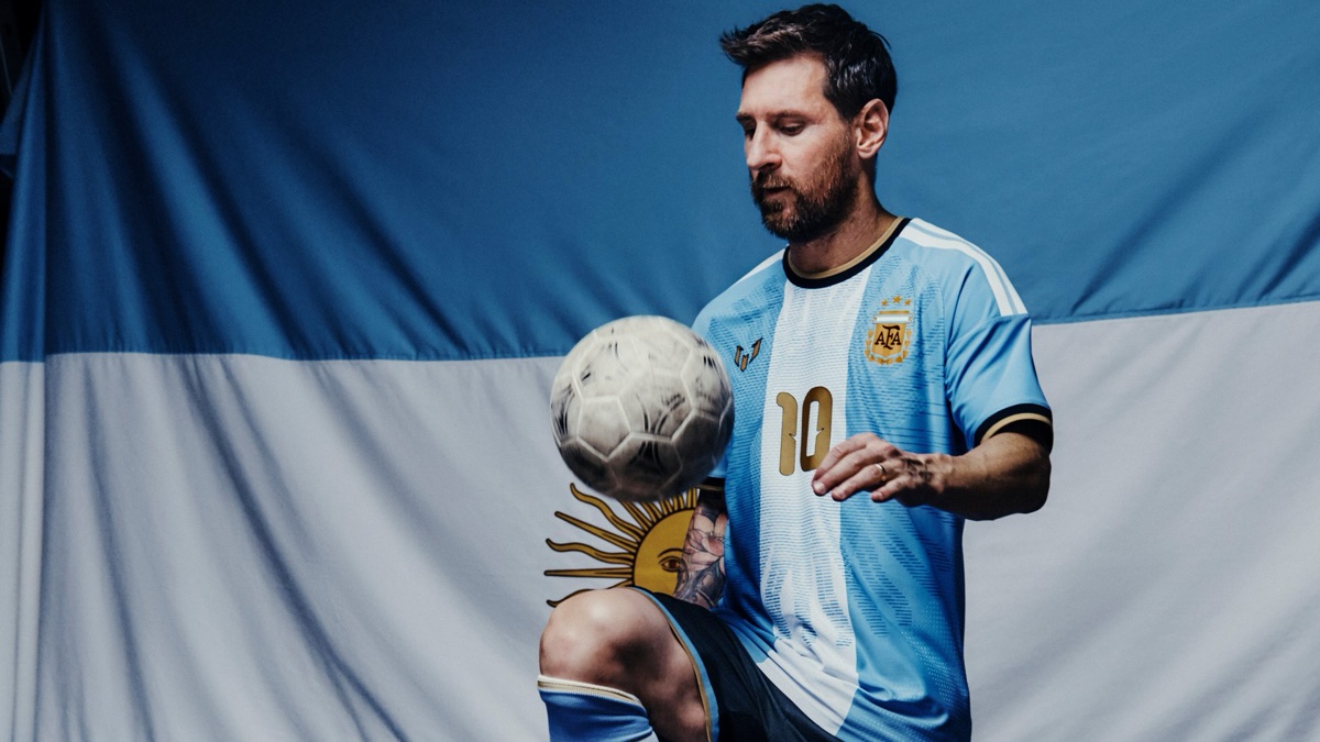 Lionel Messi hace jueguitos con una pelota vestido con la camiseta especial de su despedida, con el emblema personal en el pecho y el 10 dorado, frente a una bandera argentina (foto oficial de adidas)