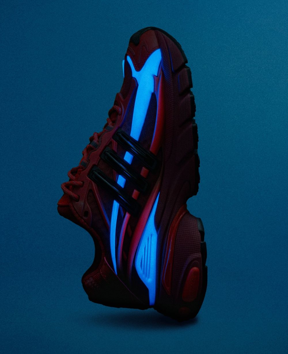 Pharrell x adidas Adistar Jellyfish Red de perfil sobre fondo azul oscuro, con las capas laterales que brillan en celeste en la oscuridad sobre la base roja