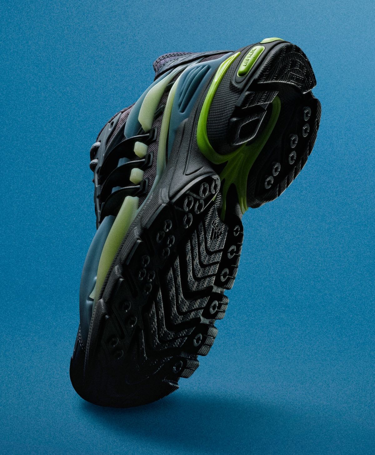 Suela del Pharrell x adidas Adistar Jellyfish Storm vista desde abajo sobre fondo azul, con tacos de goma negros, capas grises y la mediasuela con detalles verde claro y volt