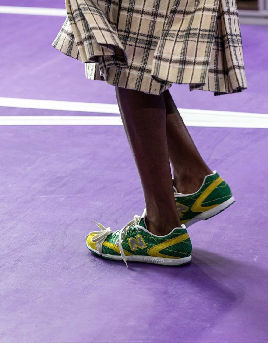Foto oficial del desfile Primavera/Verano 2027 de Miu Miu: New Balance RC150 verdes con detalles amarillos y logo N plateado, con una pollera escocesa beige, sobre la pista violeta