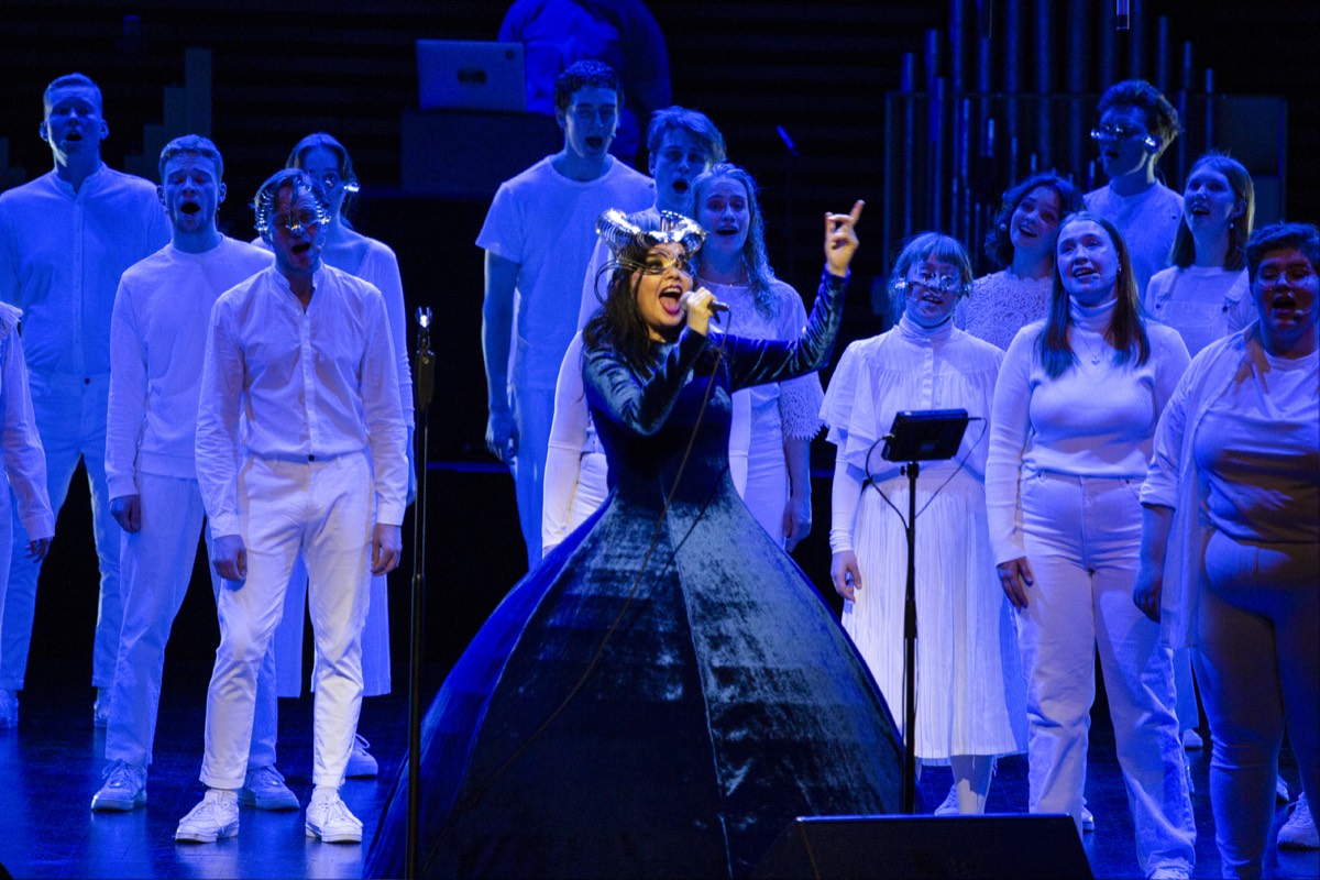 Foto oficial de Björk en el concierto Orkestral del 24 de octubre de 2021 en el Harpa de Reikiavik: con un vestido de terciopelo azul y un tocado metálico, canta con el brazo en alto delante del coro Hamrahlíð, todo vestido de blanco
