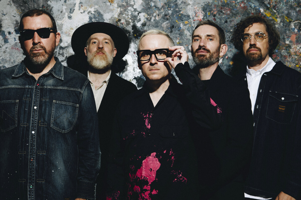 Foto de prensa de Hot Chip: los cinco integrantes de la banda vestidos de negro y de jean, parados frente a una pared manchada de pintura de colores; el del centro se acomoda unos anteojos de marco grueso negro