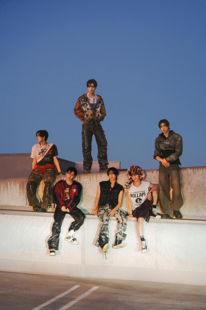 Foto de prensa oficial de JYP Entertainment de KickFlip: seis integrantes posan al atardecer sobre un muro de cemento en una terraza, tres sentados en el borde y tres parados, con una campera bomber bordó, chalecos y camperas de cuero, pantalones cargo y camuflados, zapatillas altas y un gorro de piel con estampado animal, bajo un cielo azul