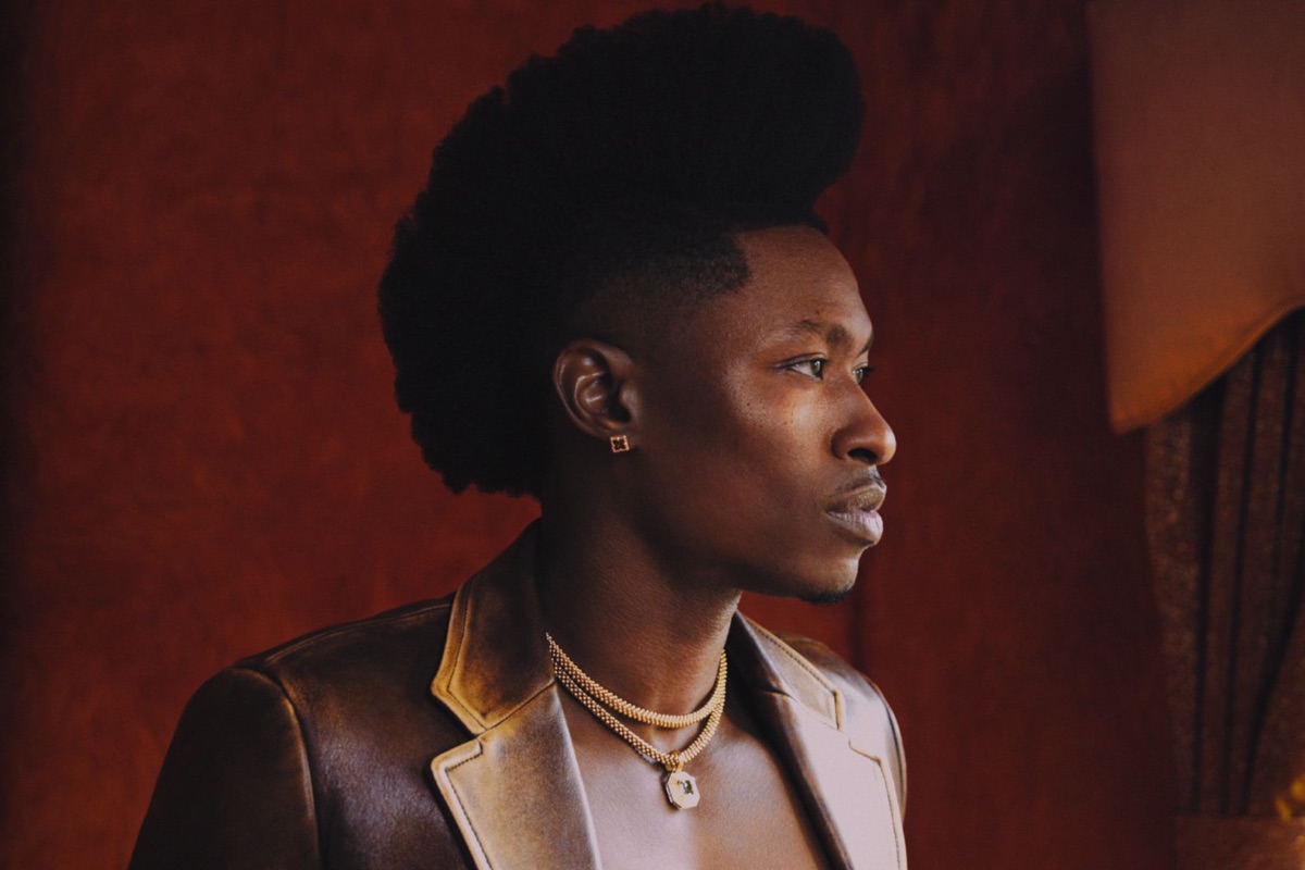 Retrato de prensa de Lucky Daye de perfil, con saco de cuero marrón, collares dorados y el pelo alto, frente a una pared bordó. Foto: Micaiah Carter