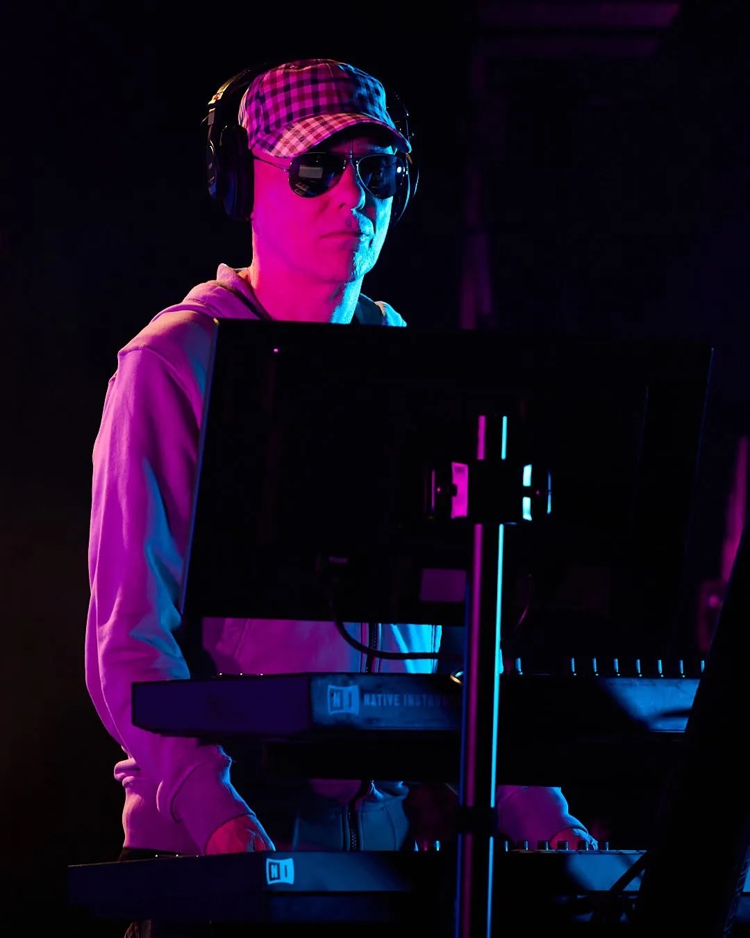 Chris Lowe tocando teclados bajo luces rosas y azules, con gorra a cuadros, anteojos oscuros, auriculares y buzo claro; foto publicada por Pet Shop Boys
