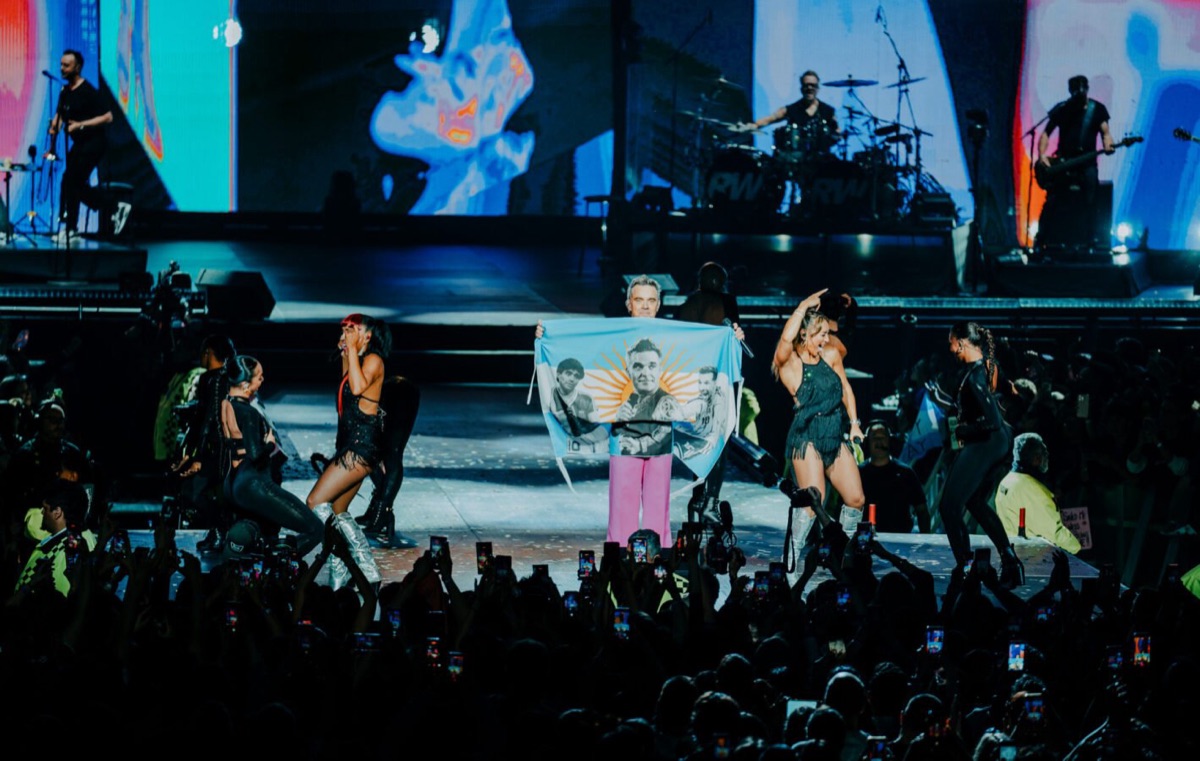 Robbie Williams con pantalón rosa sostiene una bandera argentina con su cara y el sol estampados, rodeado de bailarinas y con la banda detrás, en uno de sus shows de este mes en el Movistar Arena de Buenos Aires