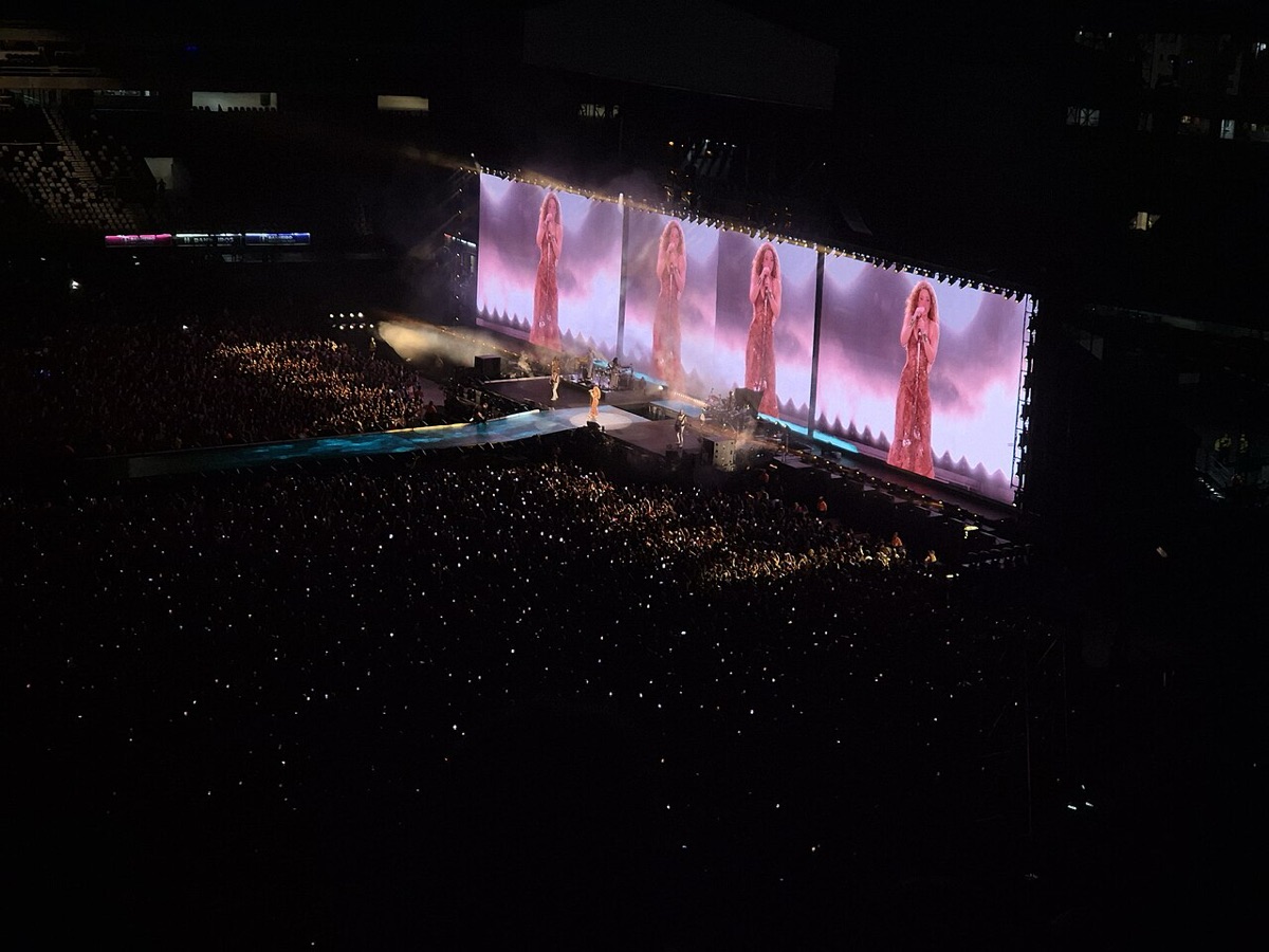 Foto de archivo de un show de la gira Las Mujeres Ya No Lloran: un estadio a oscuras con el público iluminado por celulares y una pantalla gigante que muestra a Shakira con vestido rojo, en Río de Janeiro, en 2025. Foto: Priscila Bruder, Wikimedia Commons, CC0
