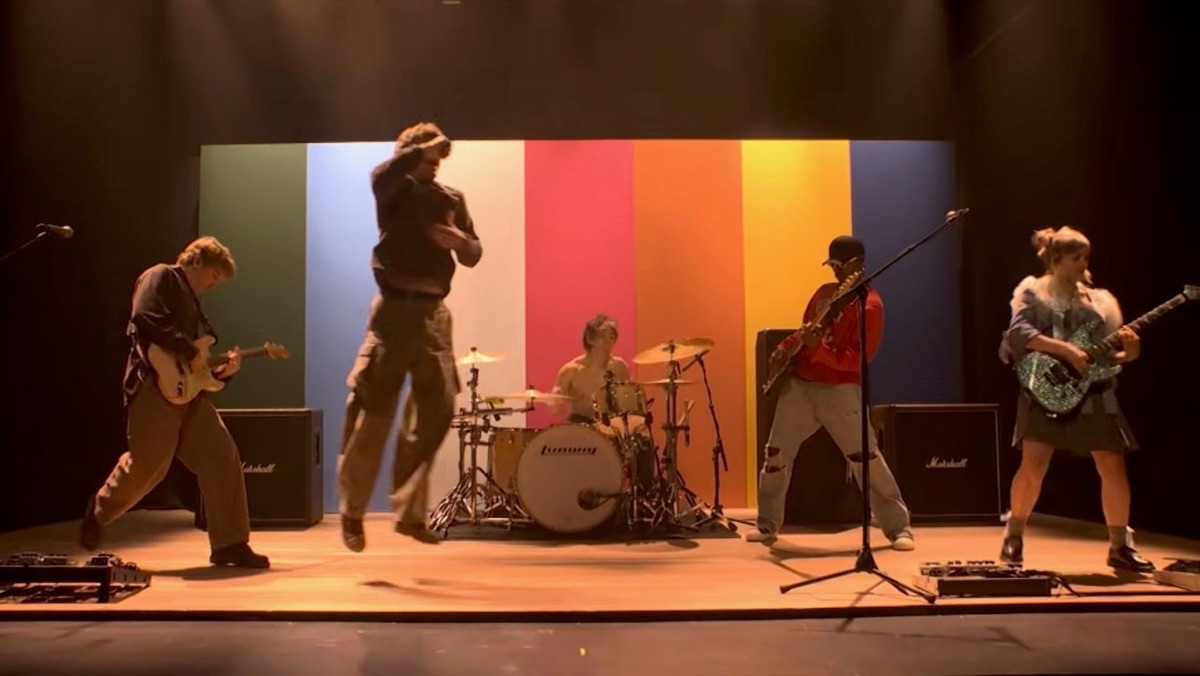 Los cinco integrantes de Turnstile tocando sobre un escenario de madera frente a un fondo de franjas verticales verde, celeste, blanca, roja, naranja, amarilla y azul; el cantante salta en el aire y el baterista toca sin remera en el centro