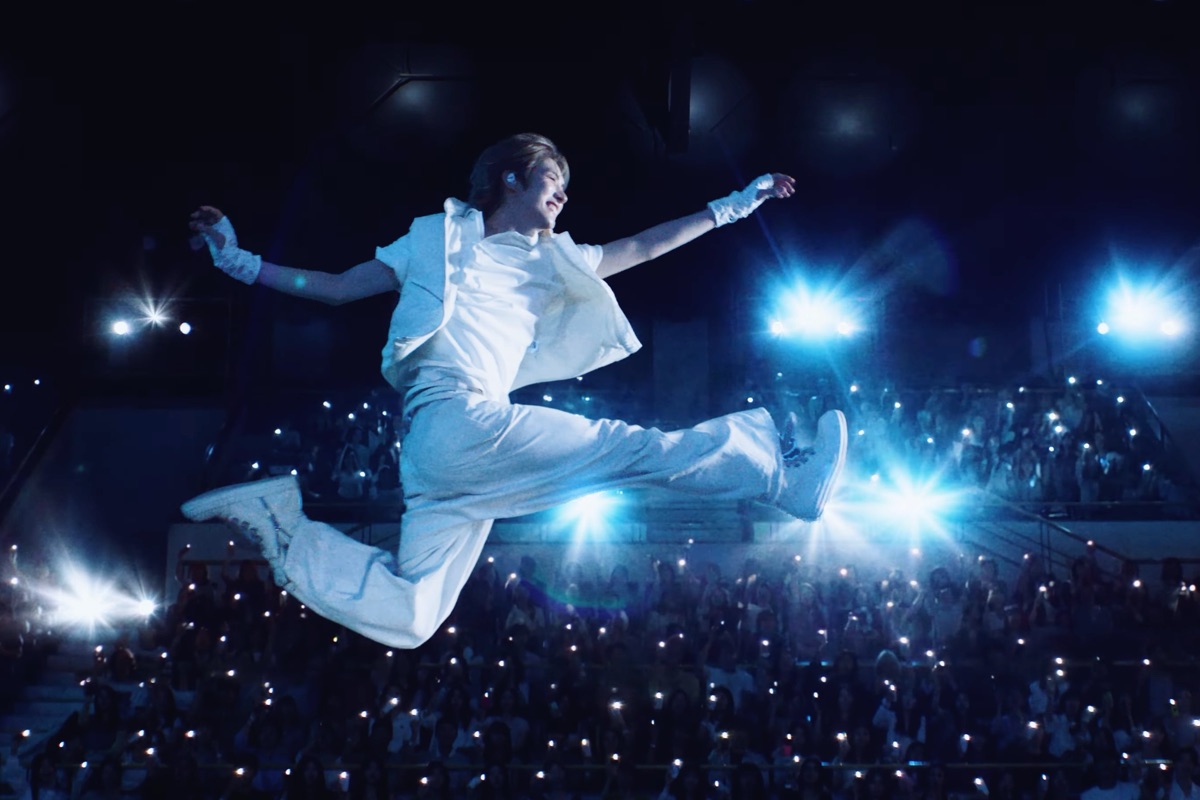 Imagen oficial del tráiler "Being Together : TO US": un integrante de TWS, vestido de blanco, salta con las piernas abiertas sobre un estadio lleno de luces de celulares