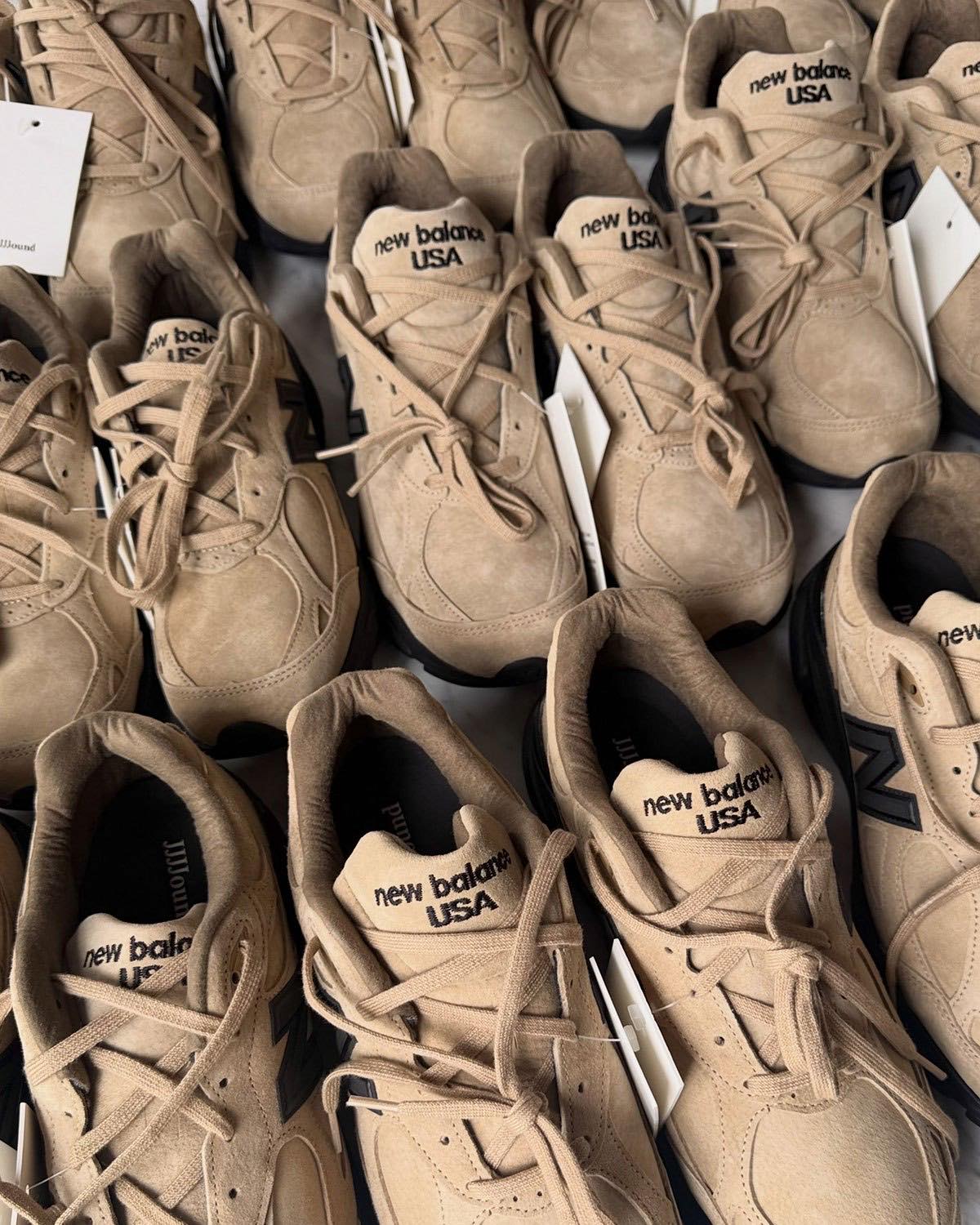 Varios pares del JJJJound x New Balance 990v3 Tan Suede apilados, con etiquetas colgantes blancas y las lengüetas new balance USA a la vista