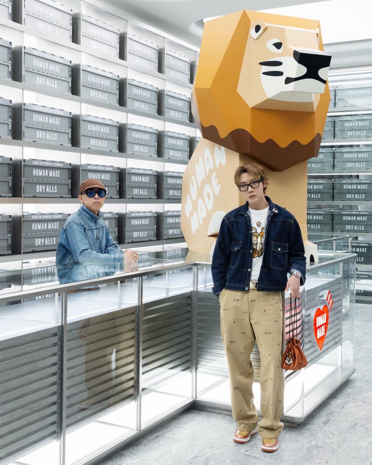 NIGO apoyado en un mostrador de vidrio y j-hope de pie delante de una gran escultura de león de Human Made, con el Air Force 1 Burnt Sunrise en los pies