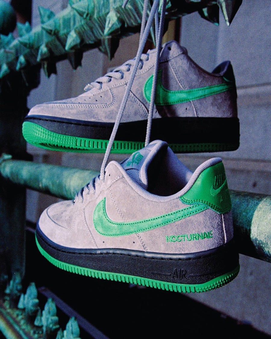Foto de campaña de dos Nike SB Air Force 1 de Nocturnal colgadas de los cordones de una reja verde con puntas, con upper de gamuza gris, Swoosh y talón verdes, mediasuela negra, suela verde y la palabra NOCTURNAL bordada en verde en el lateral del talón