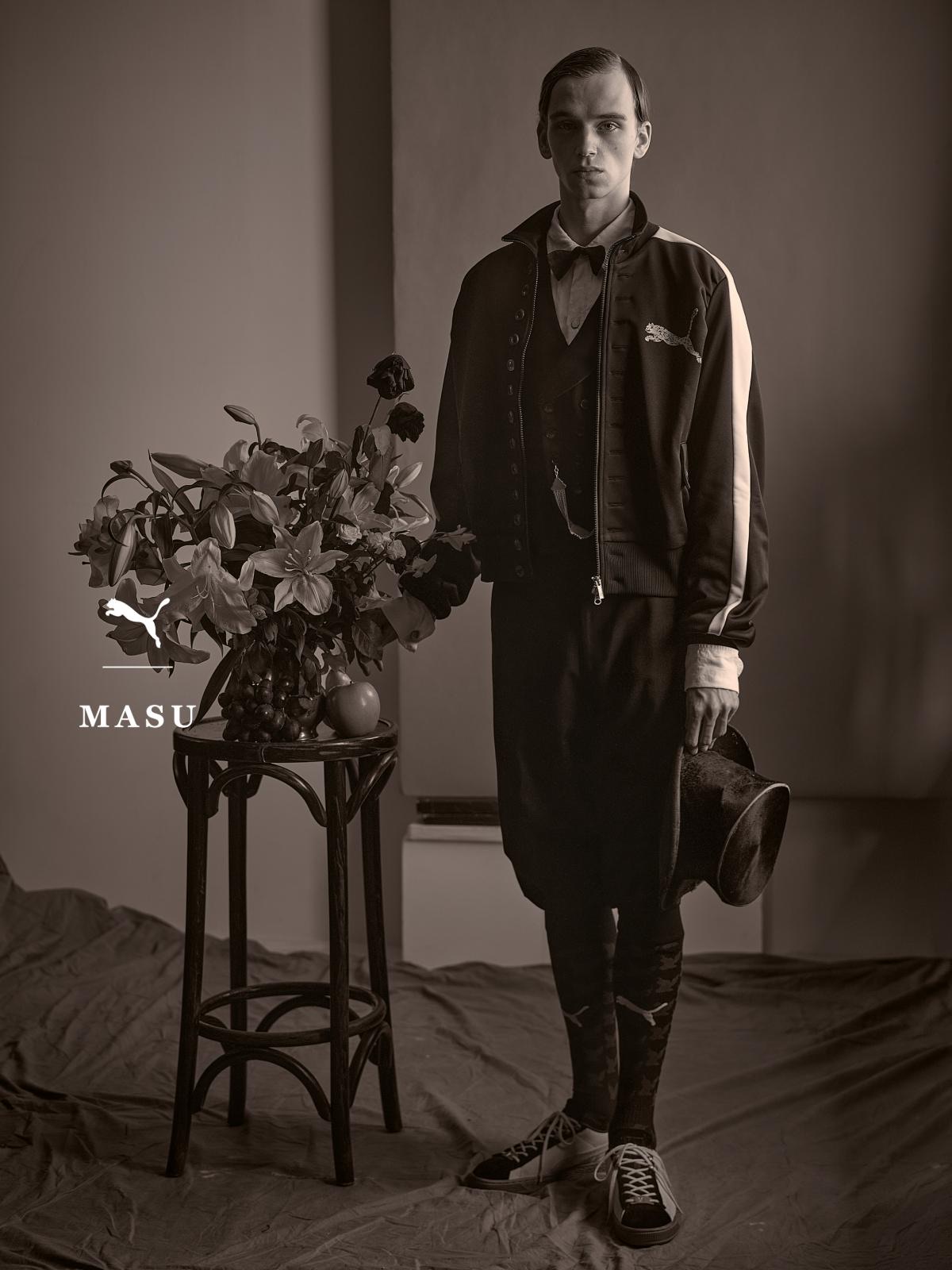 Campaña de PUMA x MASU en blanco y negro: un modelo con la campera T7 Curio negra de franjas claras, moño y sombrero en la mano, posa junto a una mesa con flores y calza las Curio Suede