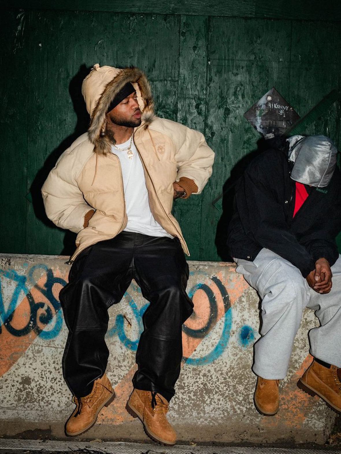 Dos modelos sentados en un murito con grafitis, uno con la campera Supreme x Timberland Suede Puffer color wheat y botas Timberland 6-Inch amarillas