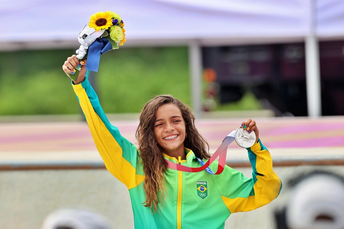 Rayssa Leal sonríe con el brazo en alto, un ramo de flores en una mano y la medalla de plata en la otra, con la campera verde y amarilla de Brasil