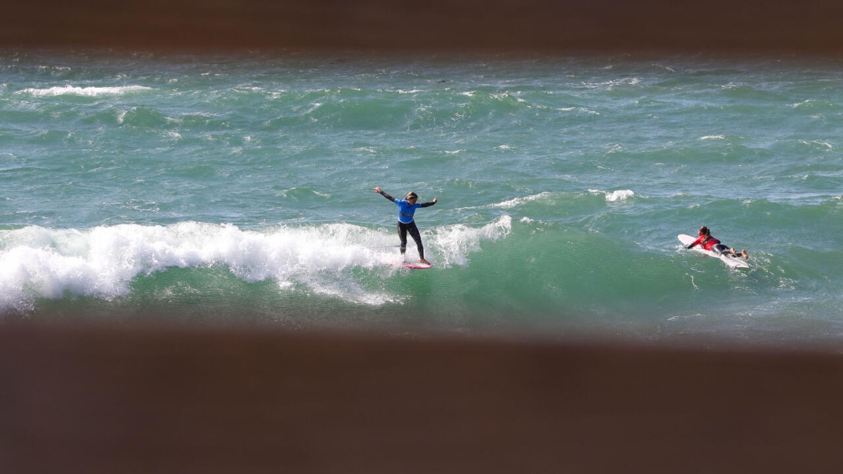 Story Martinez, con lycra azul, abre los brazos parada sobre su longboard en la espuma de una ola de Pismo Beach
