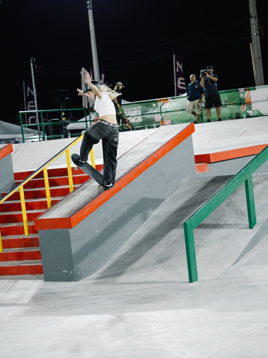 Daniela Terol, con casco, top blanco y pantalón negro, se desliza sobre el borde de un cajón rojo junto a la escalera de la pista de street de Luque, de noche, con dos camarógrafos detrás